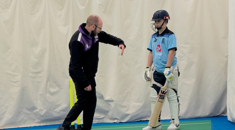 Coach observing a batter during a one-to-one cricket coaching session in outdoor nets.