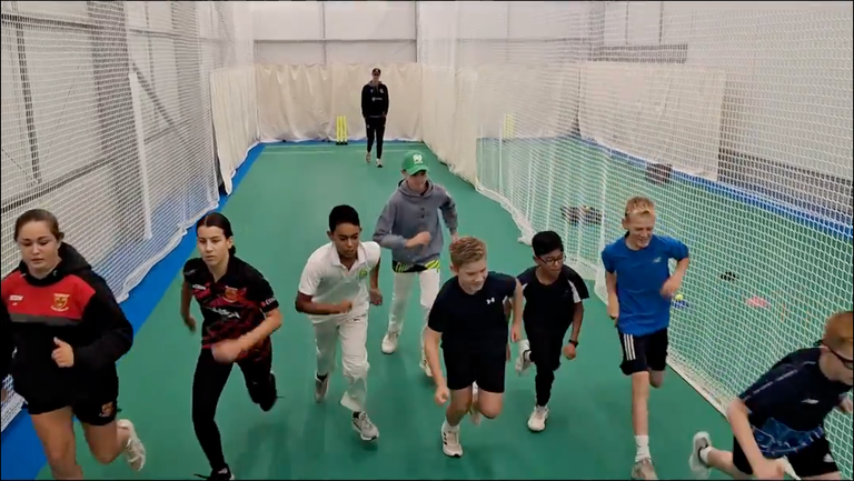Young cricket players running together during a training session inside an indoor cricket net facility, with a coach observing in the backgr